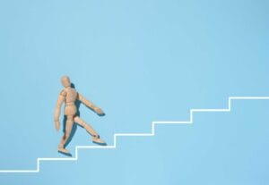 A wooden mannequin goes up a painted white staircase on a blue background. The concept of achieving the goal, overcoming obstacles