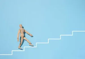 A wooden mannequin goes up a painted white staircase on a blue background. The concept of achieving the goal, overcoming obstacles